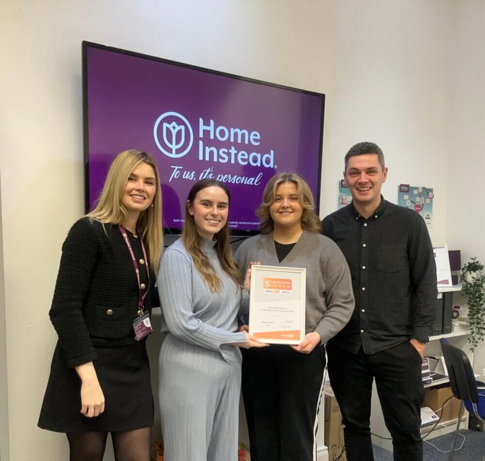 Four people smiling and holding an award in front of a screen displaying the Home Instead logo. - Home Instead