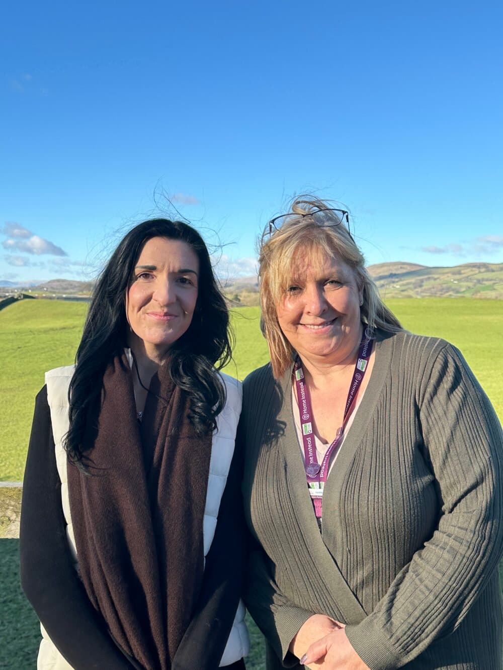 Two women standing together outside on a sunny day, with rolling green hills and a clear blue sky in the background. - Home Instead