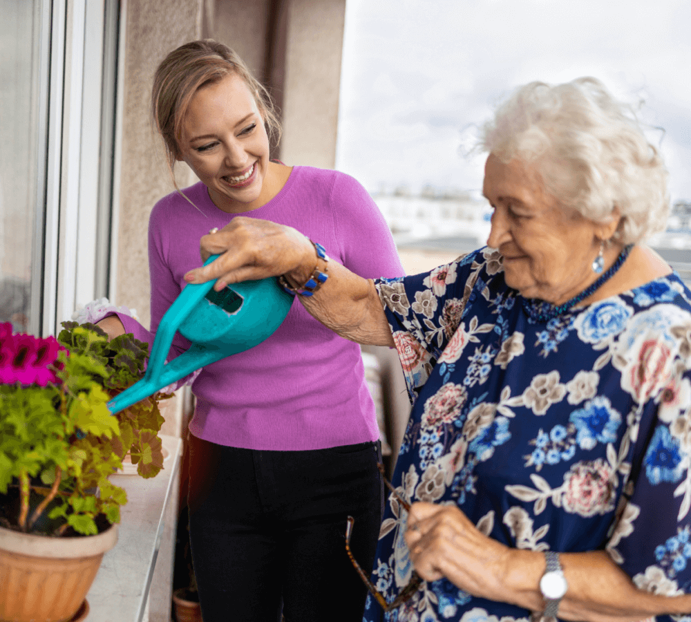 Two women, one elderly and one young, smiling while watering potted flowers on a balcony. - Home Instead