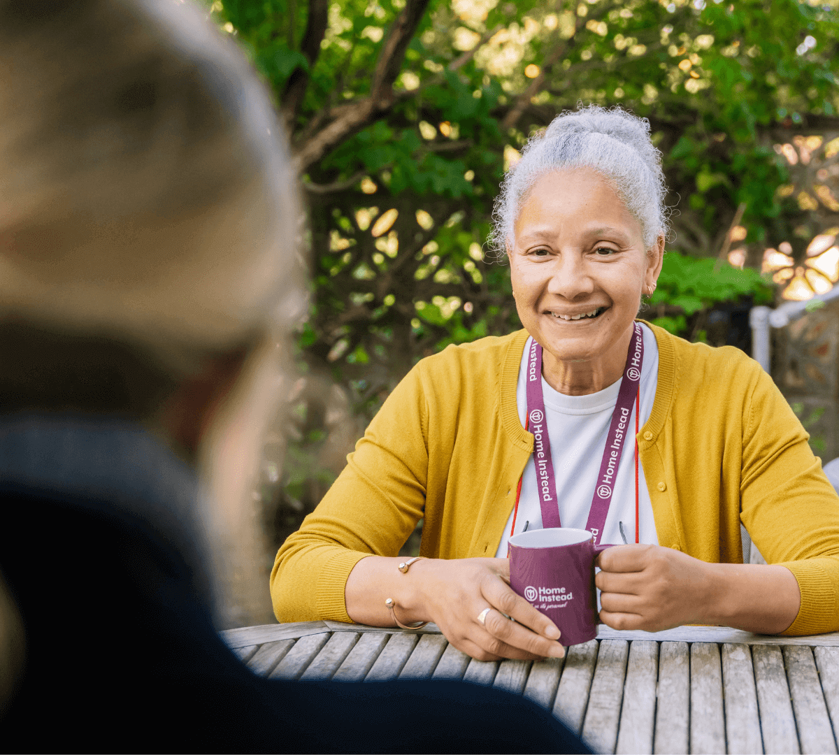 A woman with a name tag sits at a table holding a mug, smiling at another person outdoors. - Home Instead
