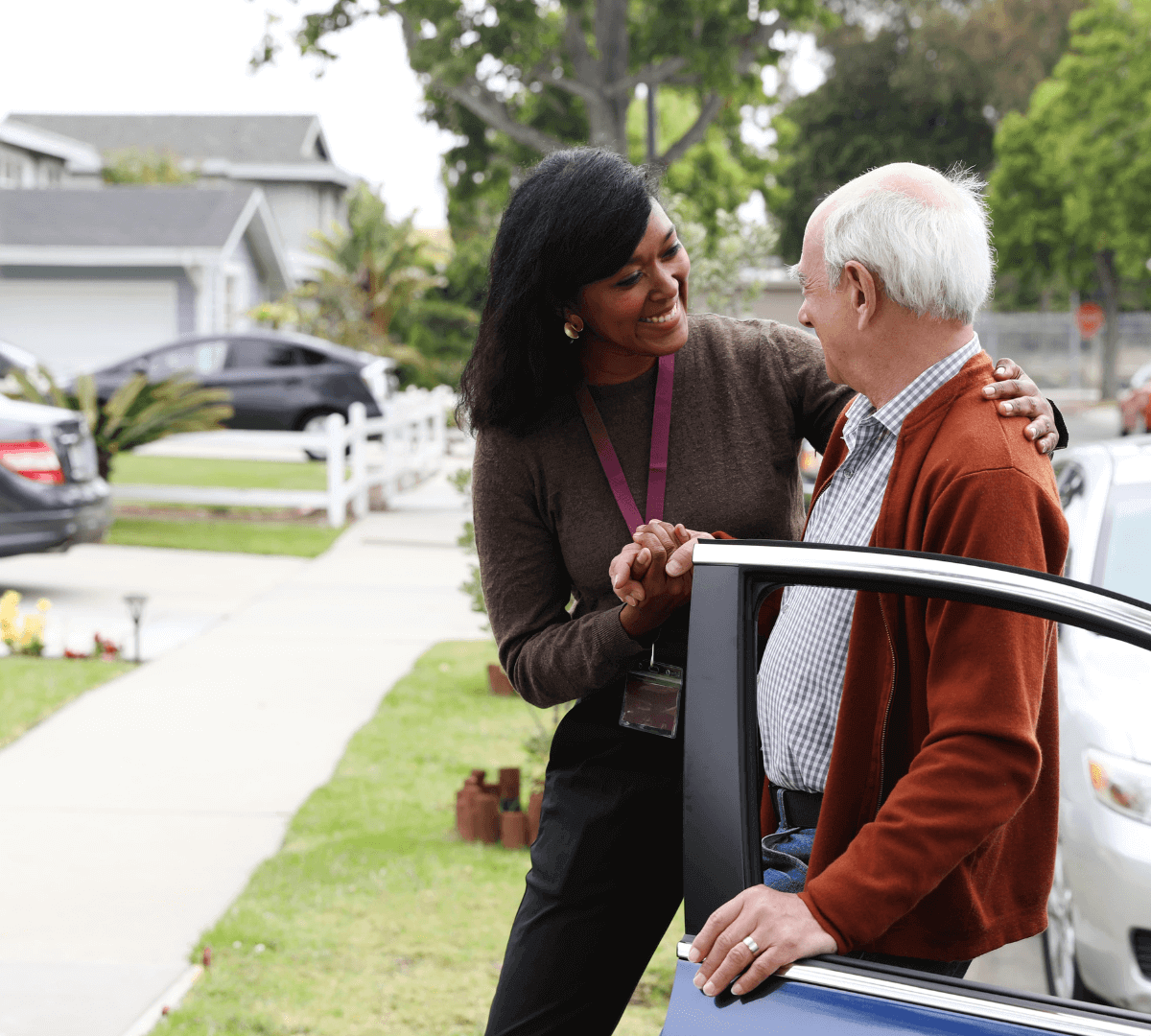 A caregiver helps an elderly man out of a car on a suburban street, both smiling warmly at each other. - Home Instead