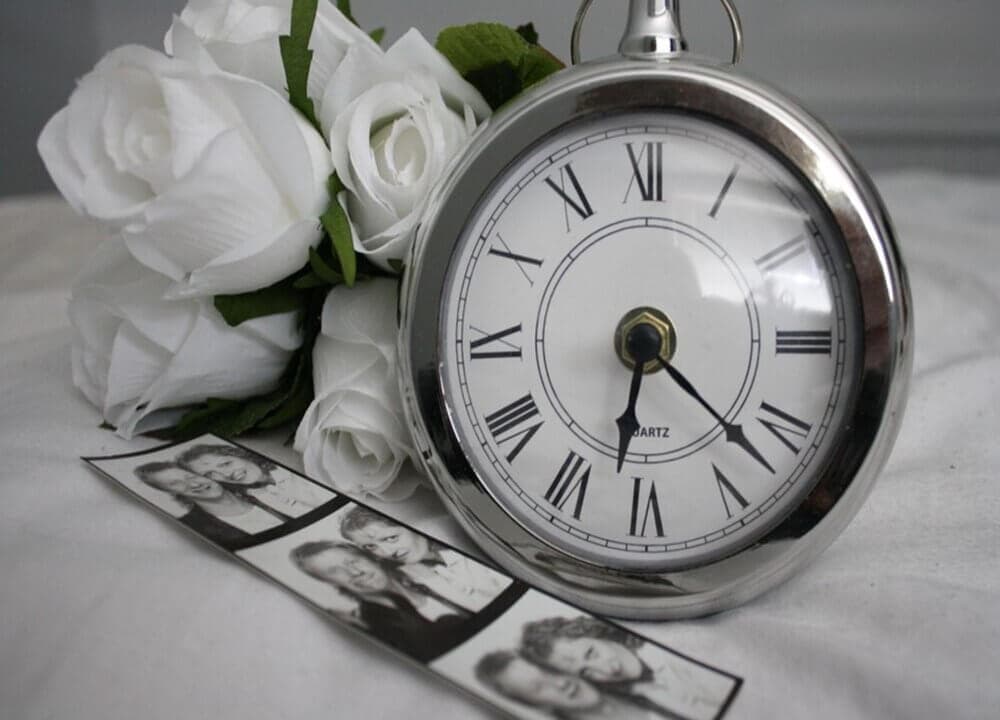 Black-and-white clock next to white roses and a strip of photo booth pictures on a white surface. - Home Instead