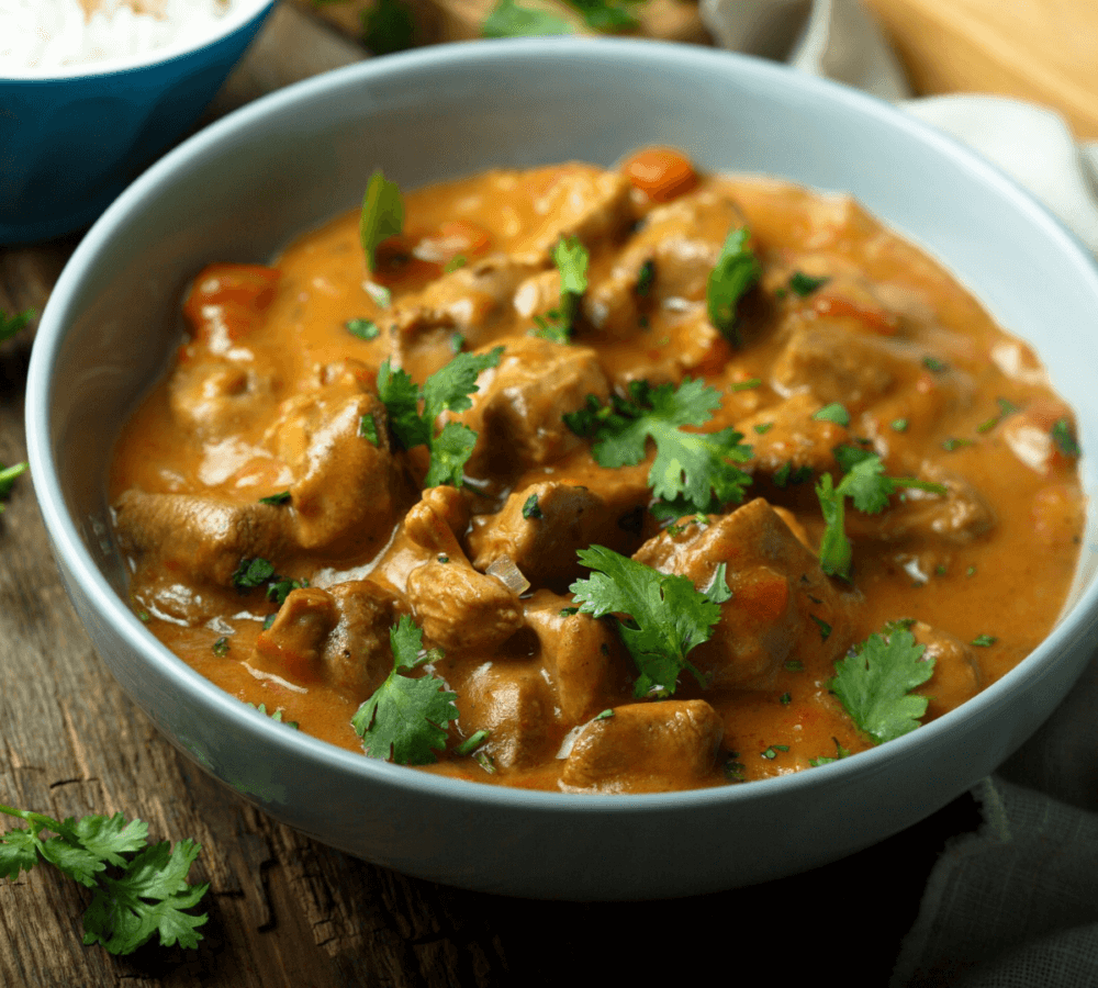 A bowl of creamy chicken curry garnished with fresh cilantro, served on a wooden table. - Home Instead