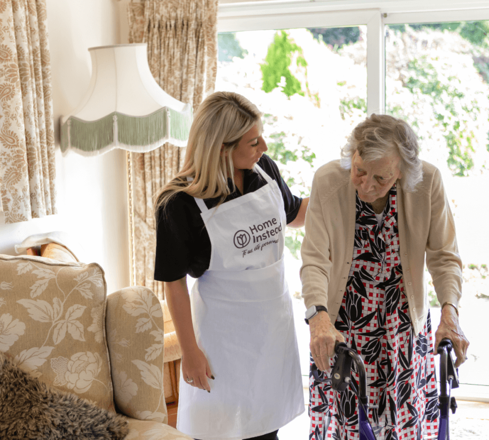 A caregiver assists an elderly woman using a walker in a cozy living room with floral decor and a large window. - Home Instead