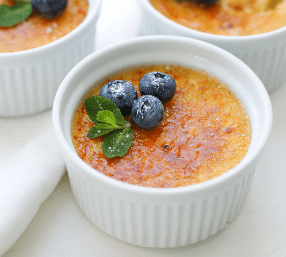 Three ramekins of crème brûlée topped with blueberries and mint leaves on a white surface. - Home Instead
