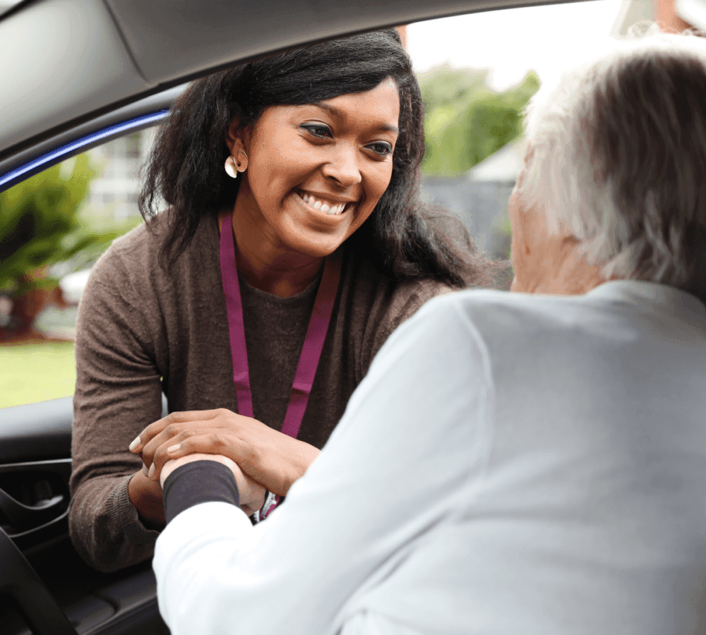A woman smiling and shaking hands with an elderly person sitting in a car. - Home Instead