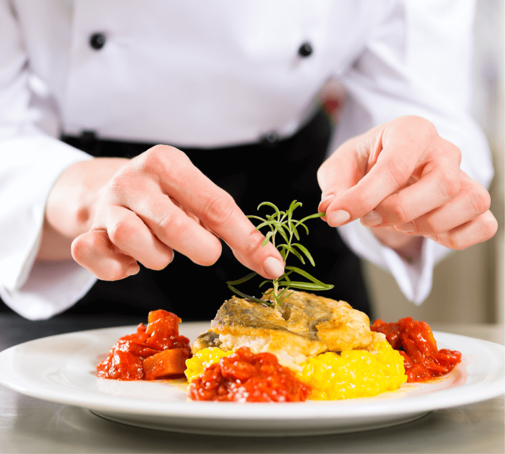 Chef garnishing a gourmet dish with herbs, featuring a white fish fillet, yellow risotto, and red sauce on a white plate. - Home Instead