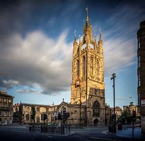 A historic cathedral with a tall, ornate tower under a blue sky with clouds, surrounded by buildings and a street. - Home Instead