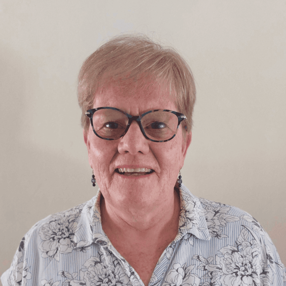 Smiling elderly person with glasses, short hair, and a patterned shirt against a plain background. - Home Instead