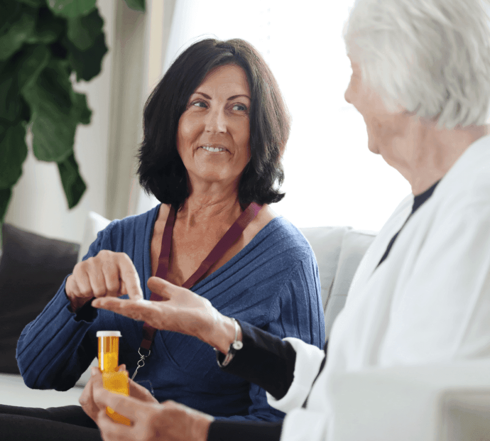 A middle-aged woman hands a pill bottle to an elderly woman at home, both smiling and seated on a couch. - Home Instead