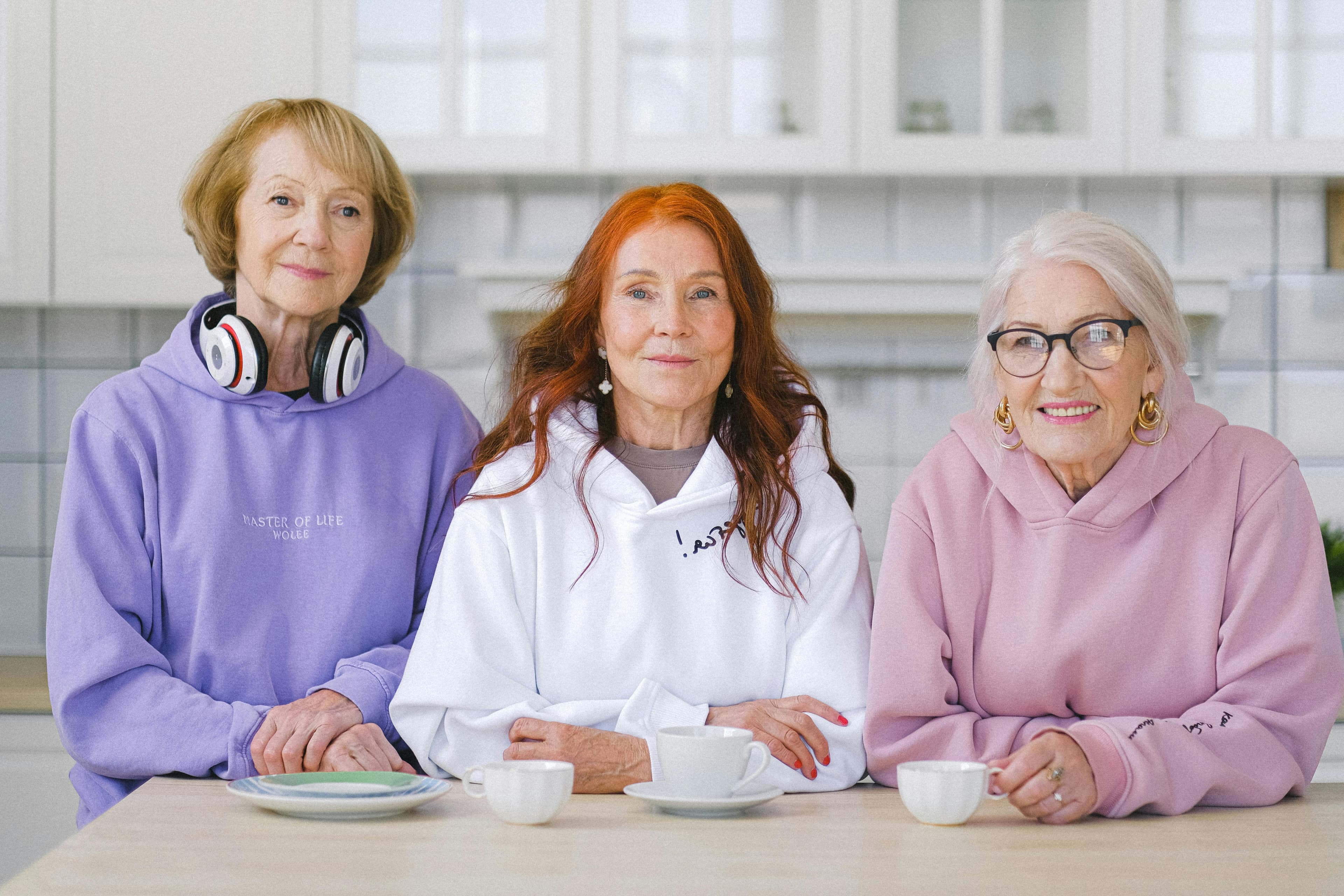 Three older women in pastel hoodies sitting at a table with cups of tea in a bright kitchen. - Home Instead
