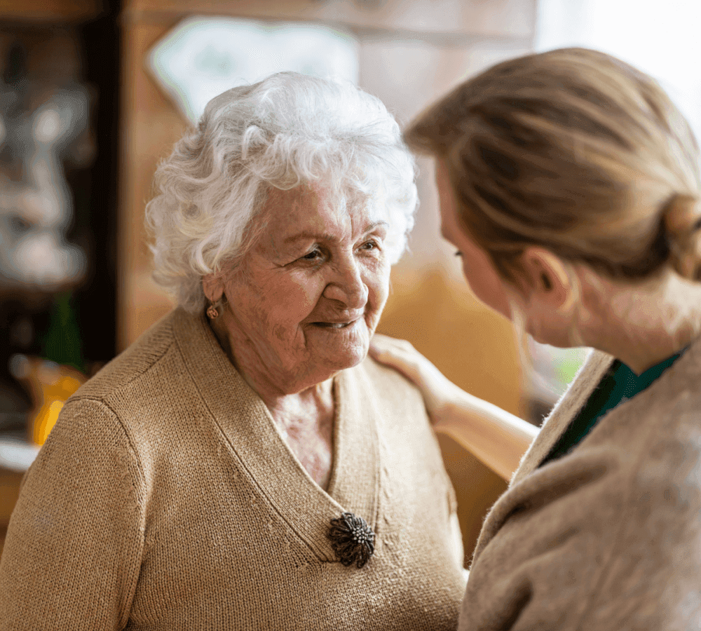 An elderly woman with white hair smiles while a younger woman with her hair tied back holds her shoulder affectionately. - Home Instead