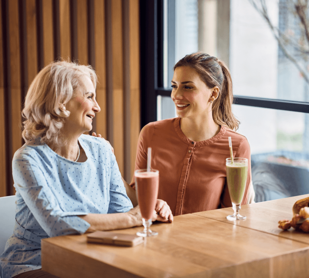 Two women seated at a table, smiling and drinking smoothies in a bright, modern café. - Home Instead