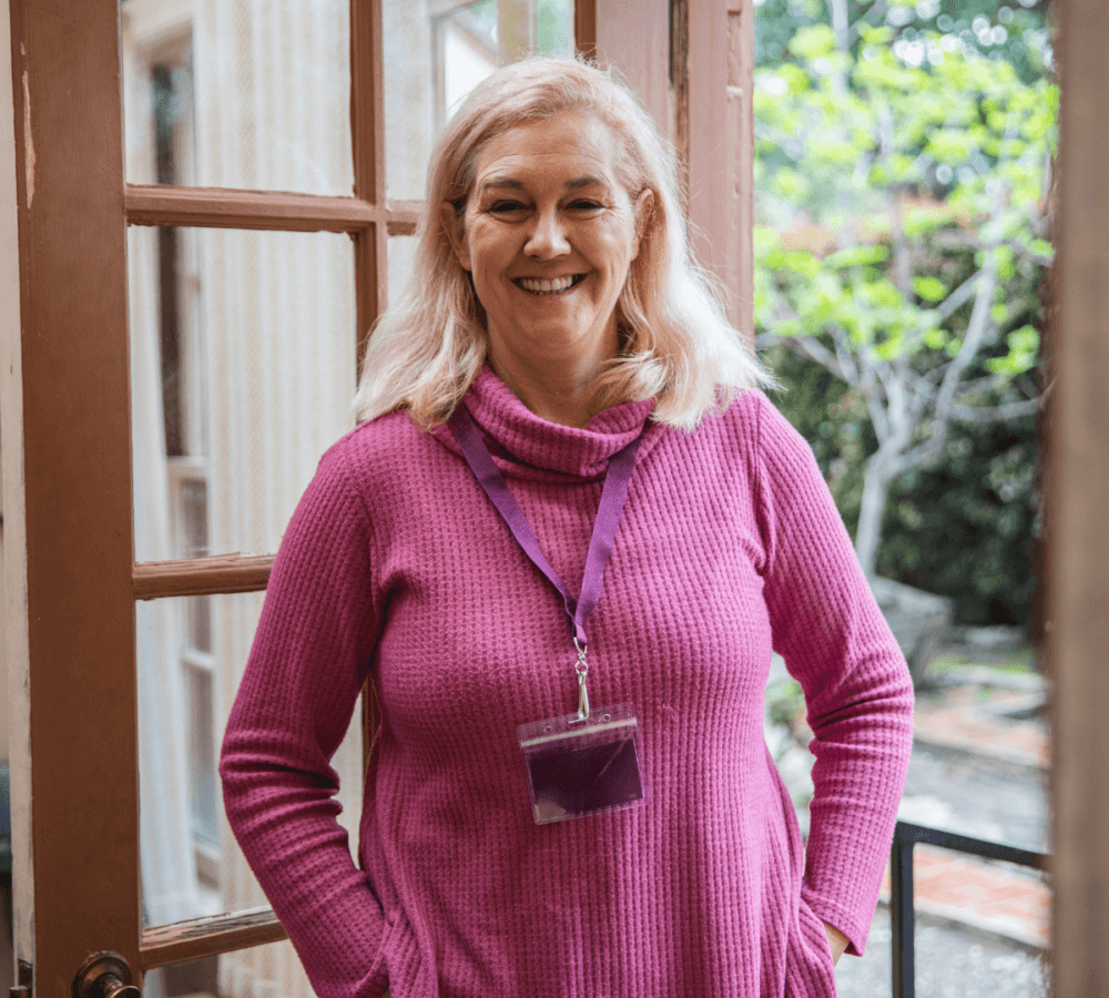 A woman with blonde hair, wearing a pink sweater and a lanyard, stands smiling in an open doorway. - Home Instead