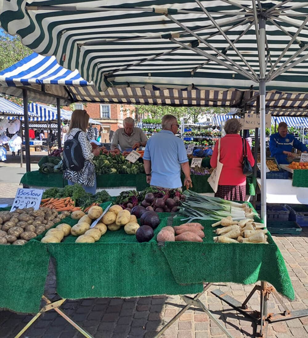 People shopping for fresh vegetables at an outdoor market under striped awnings. - Home Instead