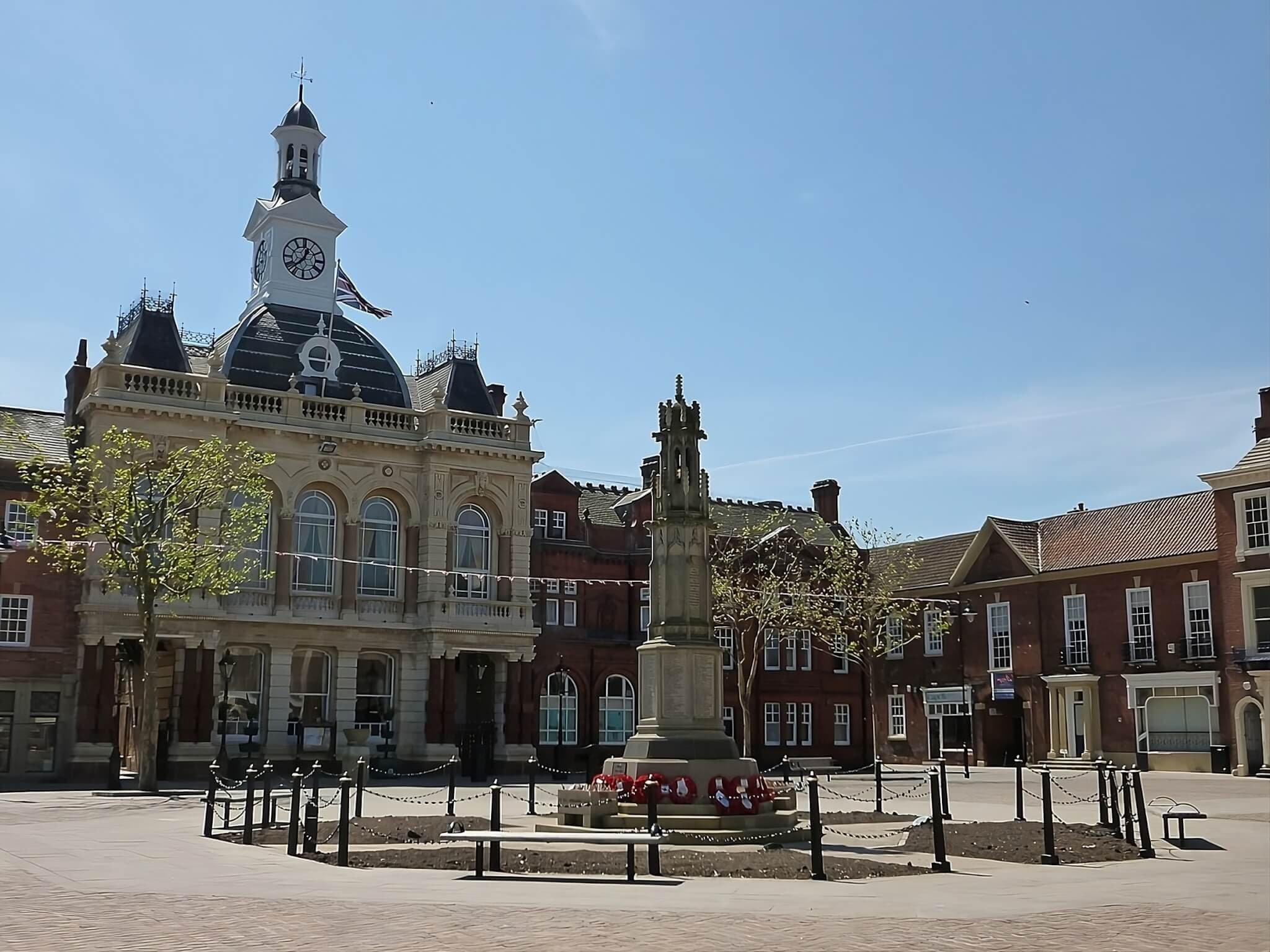 Town square with a historic building featuring a clock tower and a monument surrounded by red flowers in the foreground. - Home Instead