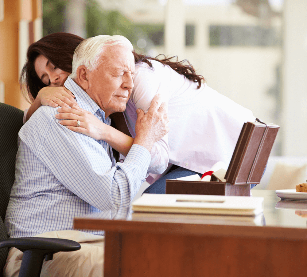 Young woman hugging an elderly man from behind while he sits at a desk with a framed photo in front of them. - Home Instead