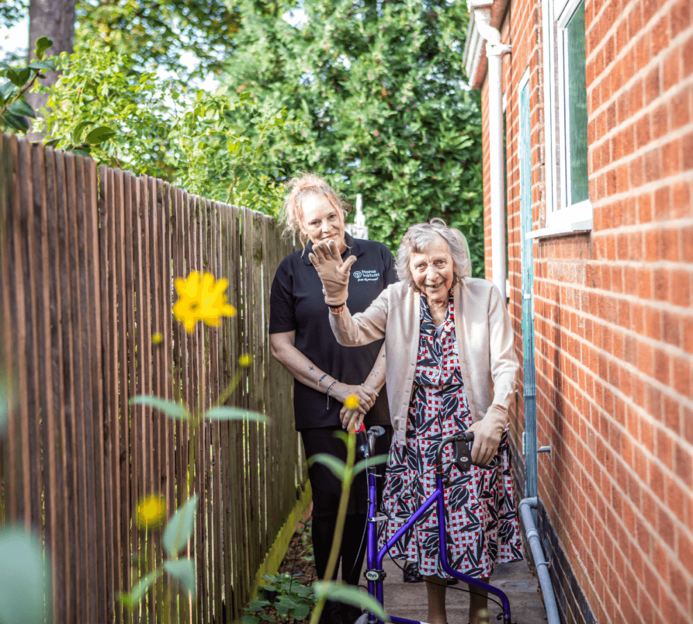 Elderly woman with walker and caretaker on a garden path, smiling and gesturing, near a brick house and wooden fence. - Home Instead