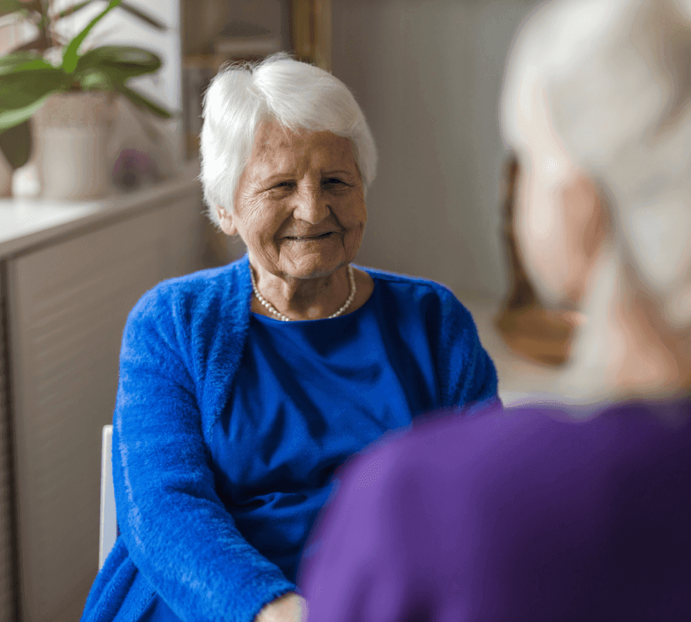 An elderly woman in a blue sweater smiles while talking to another person indoors. - Home Instead