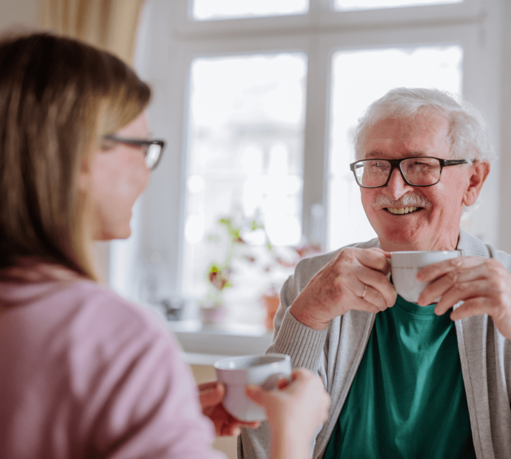 An elderly man and a younger woman smiling and enjoying coffee together in a bright room with large windows. - Home Instead