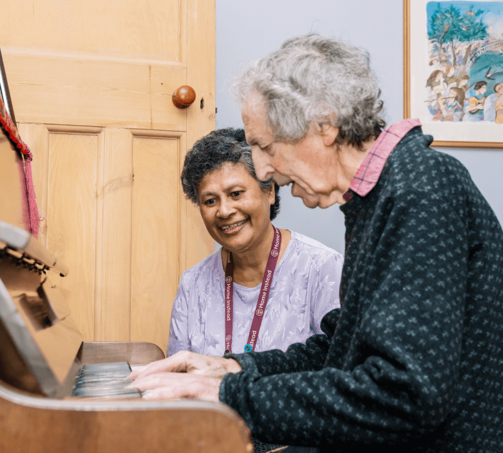 A man plays the piano while a woman watches and smiles. They are in a room with a painting and wooden door. - Home Instead