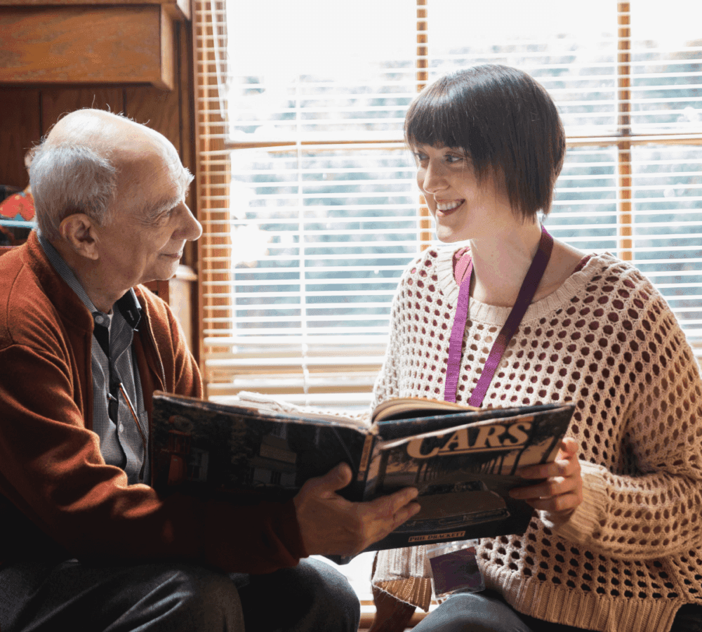 An elderly man and a young woman smile and look at a "Cars" book while sitting by a window with blinds. - Home Instead