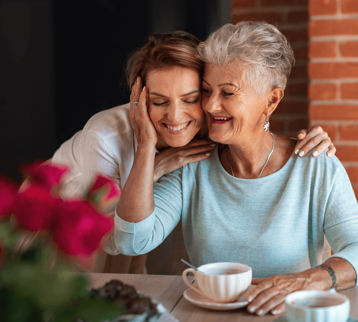 Older woman and younger woman smiling and hugging, drinking tea at a table with a vase of red flowers. - Home Instead