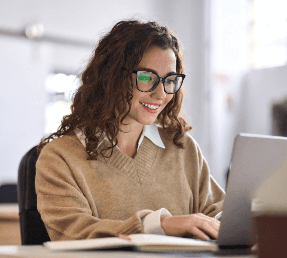 Woman with curly hair and glasses working on a laptop at a desk, smiling in a well-lit room. - Home Instead