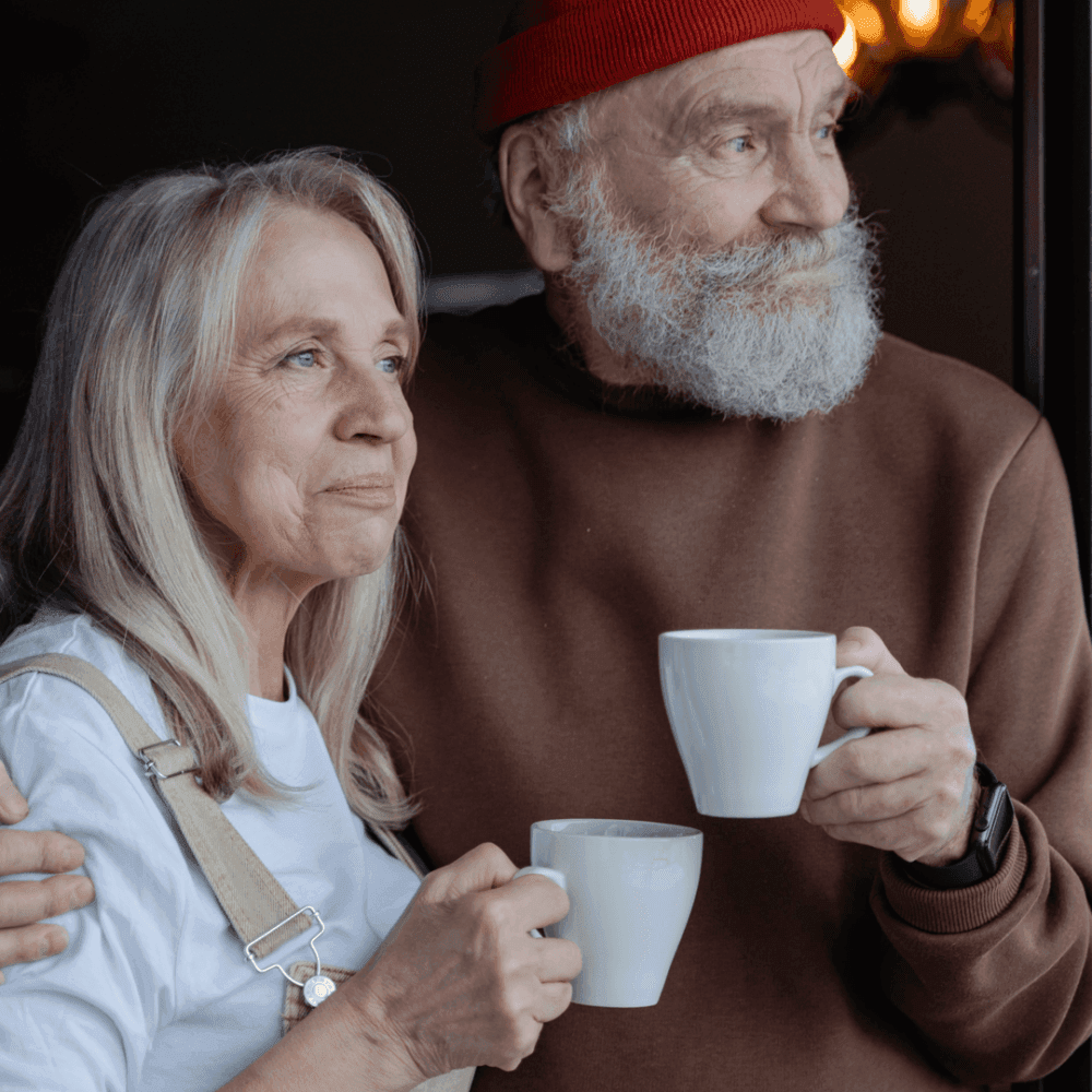 Elderly couple, woman in overalls and man with a beard in a red hat, holding white mugs and looking out the window. - Home Instead