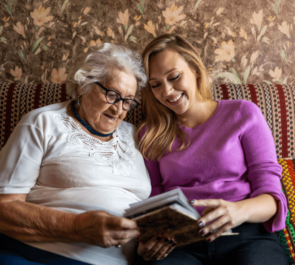 An elderly woman and a younger woman smile while looking at a photo album together on a couch. - Home Instead