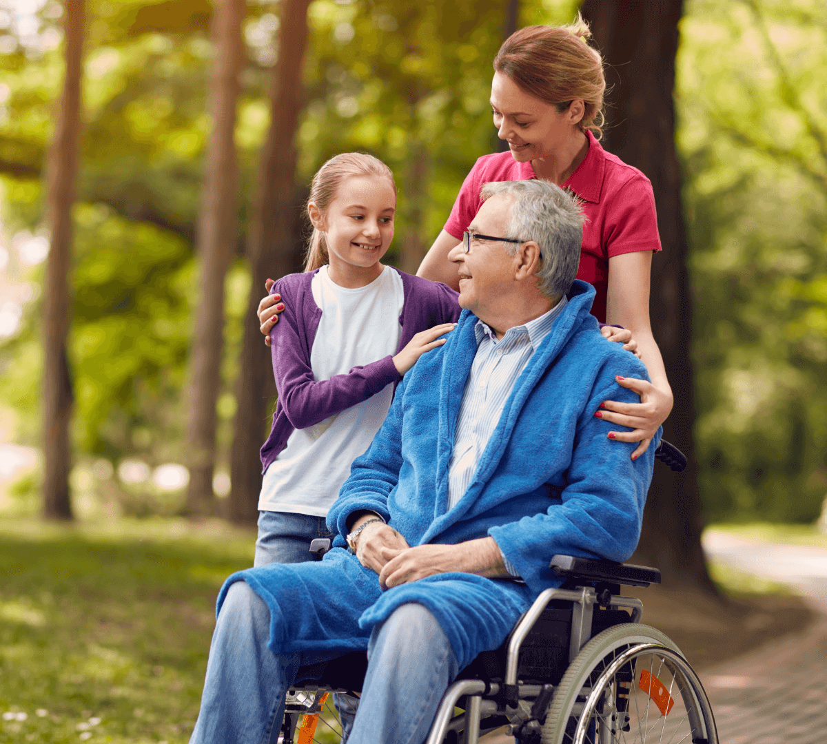 A smiling older man in a wheelchair with a young girl and a caregiver in a park on a sunny day. - Home Instead