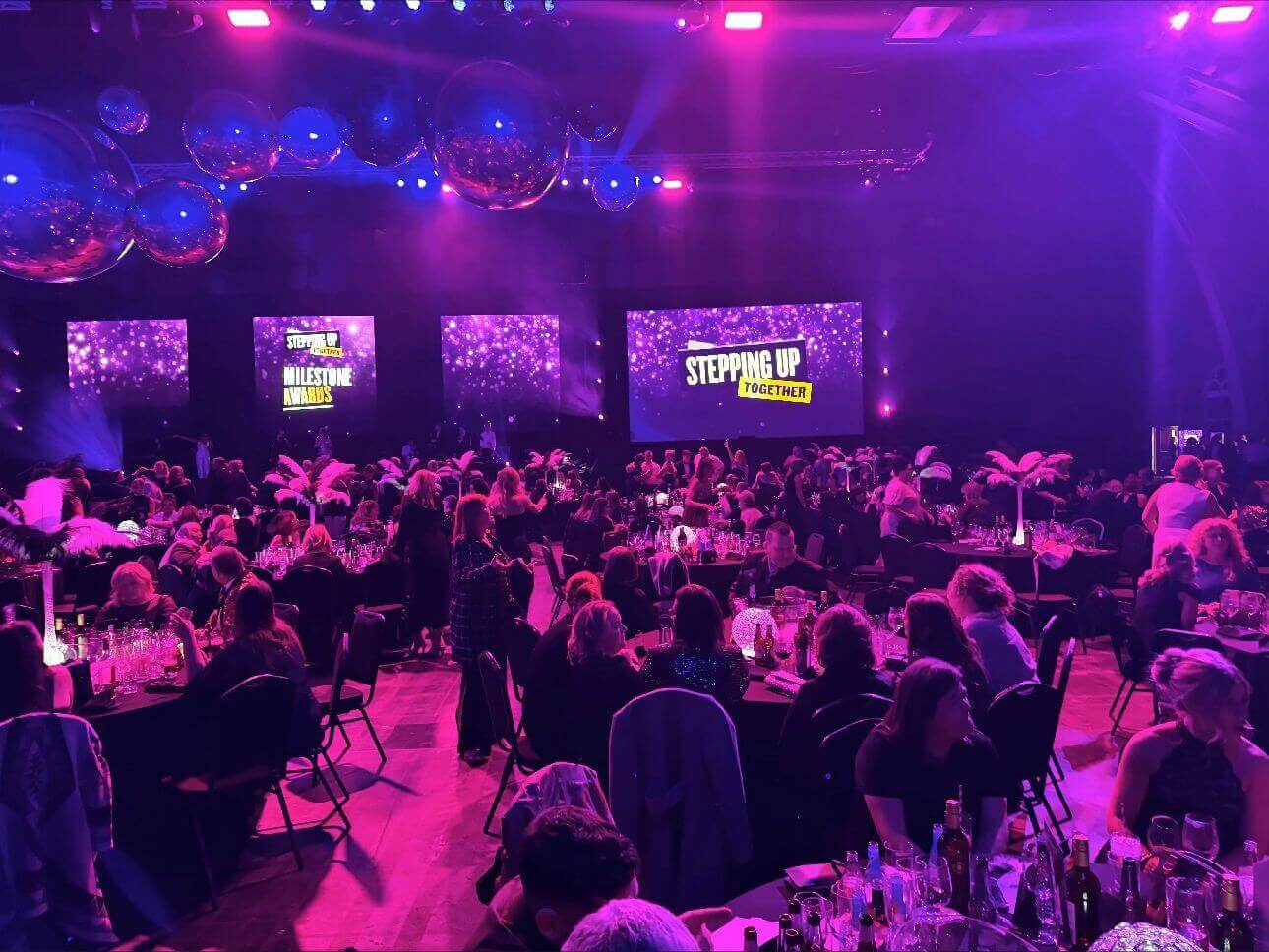 People seated at tables in a dimly lit event hall with two large screens displaying "STEPPING UP TOGETHER. - Home Instead