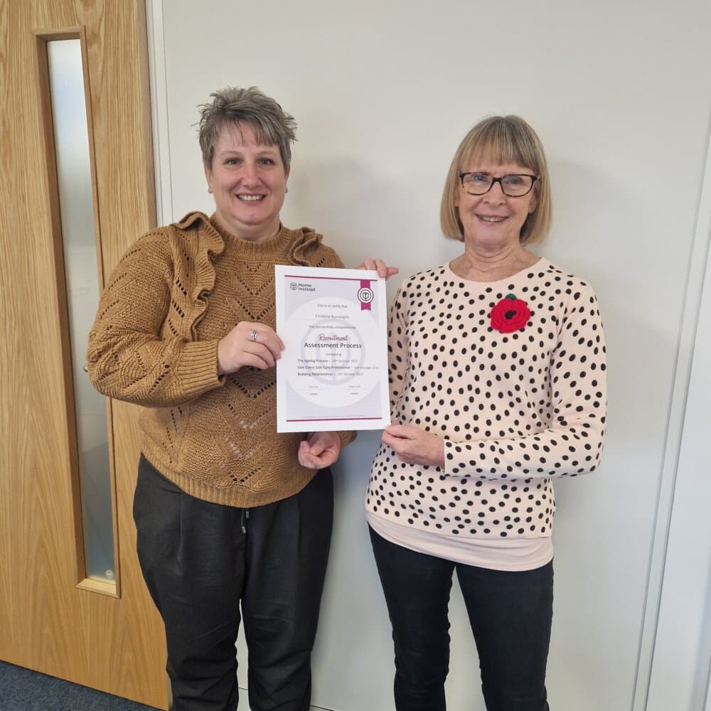 Two women smiling and holding a certificate together in an office setting. - Home Instead