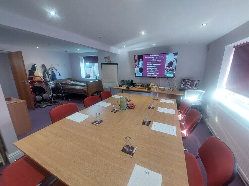 Meeting room with a large table, red chairs, presentation screen, and assorted equipment. Bed in the corner. - Home Instead