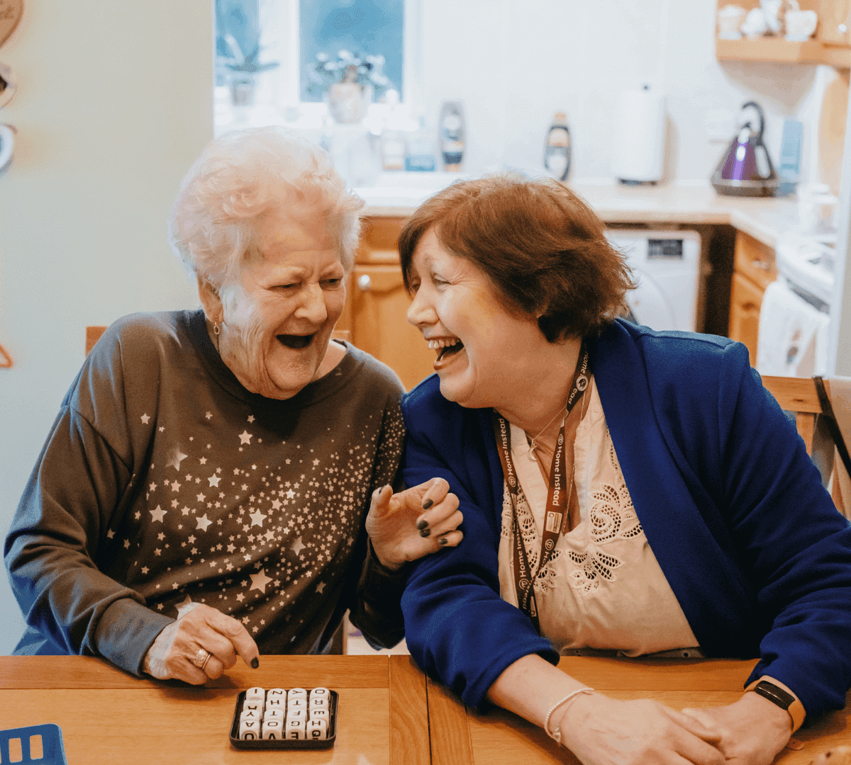 Two elderly women sitting at a table, laughing and enjoying a game together in a cozy kitchen setting. - Home Instead