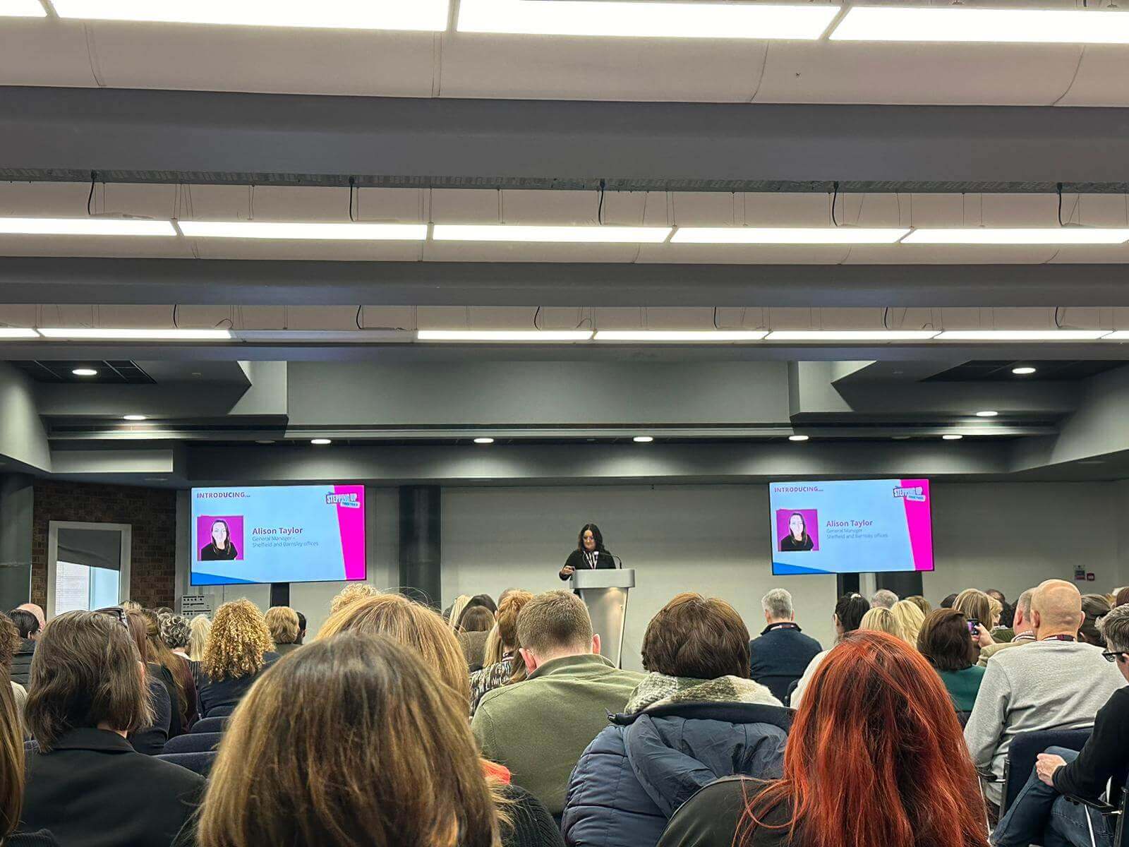 Audience seated and facing a speaker at a podium in a conference room with presentation slides displayed on screens. - Home Instead