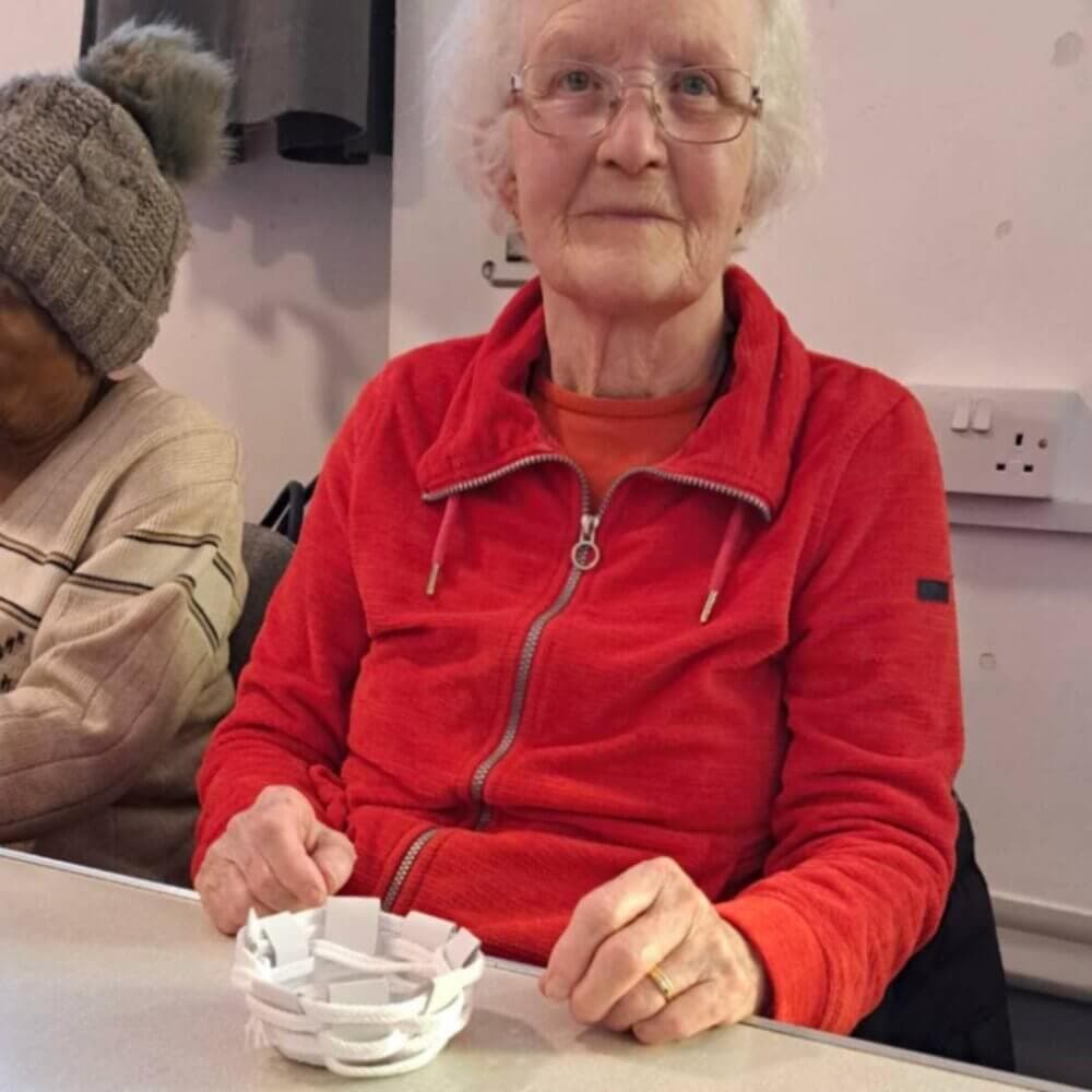 An elderly woman in a red jacket holding a small white woven basket while sitting at a table indoors. - Home Instead