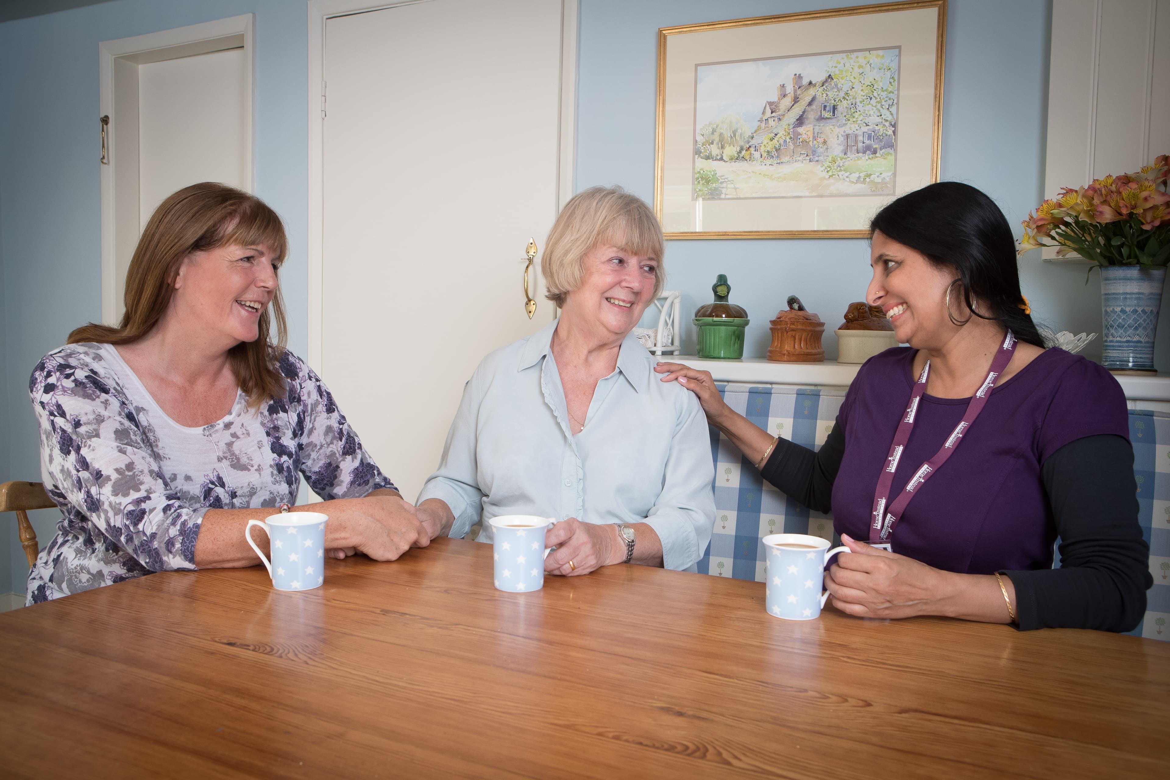 Three women sitting at a table, smiling and conversing, with mugs in front of them in a cozy room. - Home Instead
