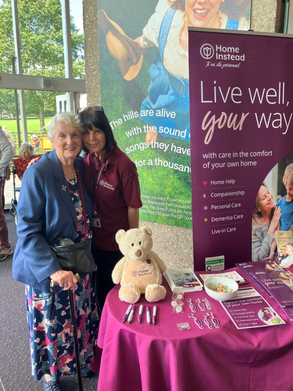 Two women smiling by a "Home Instead" information table with promotional materials and a teddy bear, in front of a display. - Home Instead