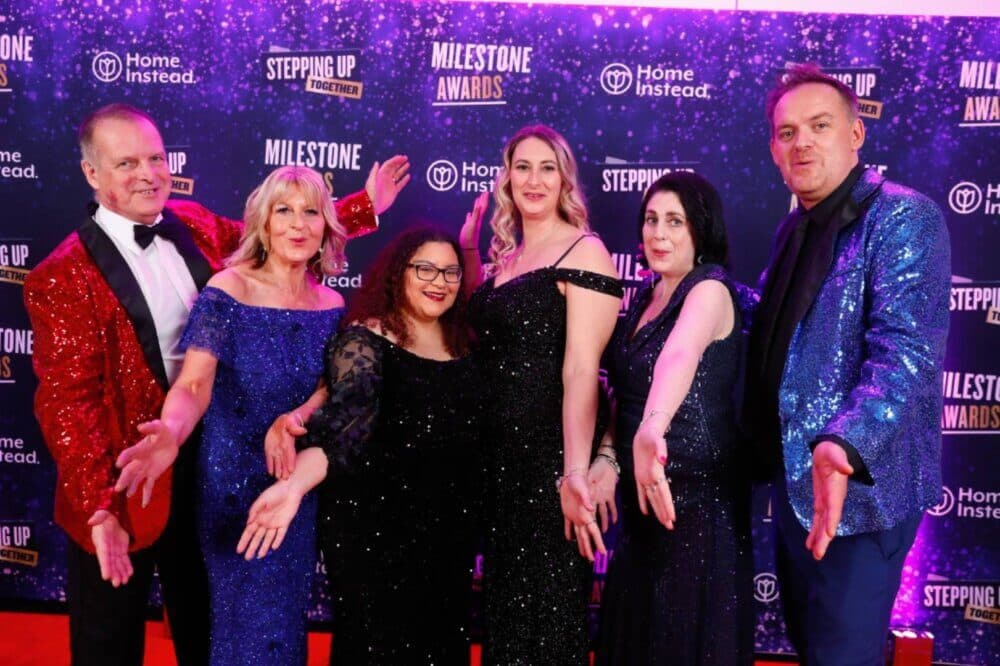 Six people in formal attire, with glittery jackets and gowns, smiling and posing on a red carpet. - Home Instead