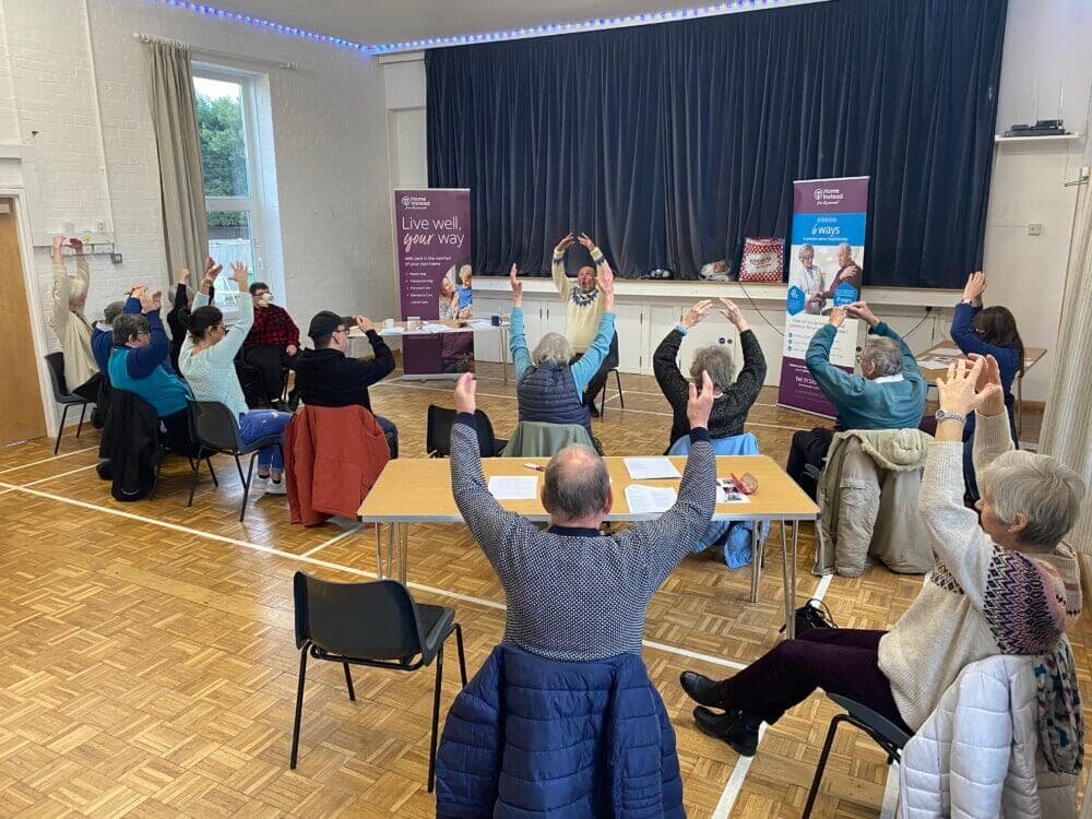 A group of seniors seated in a community hall exercising with arms raised, facing an instructor at the front. - Home Instead