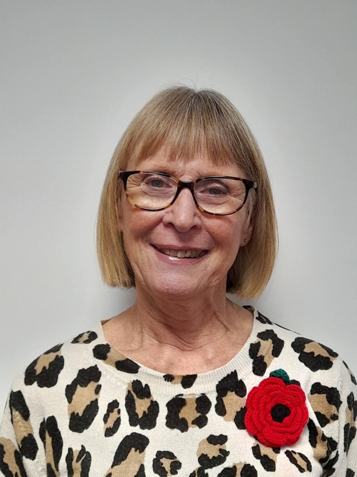Smiling woman with glasses, wearing a leopard-print top and a red poppy brooch, against a plain background. - Home Instead