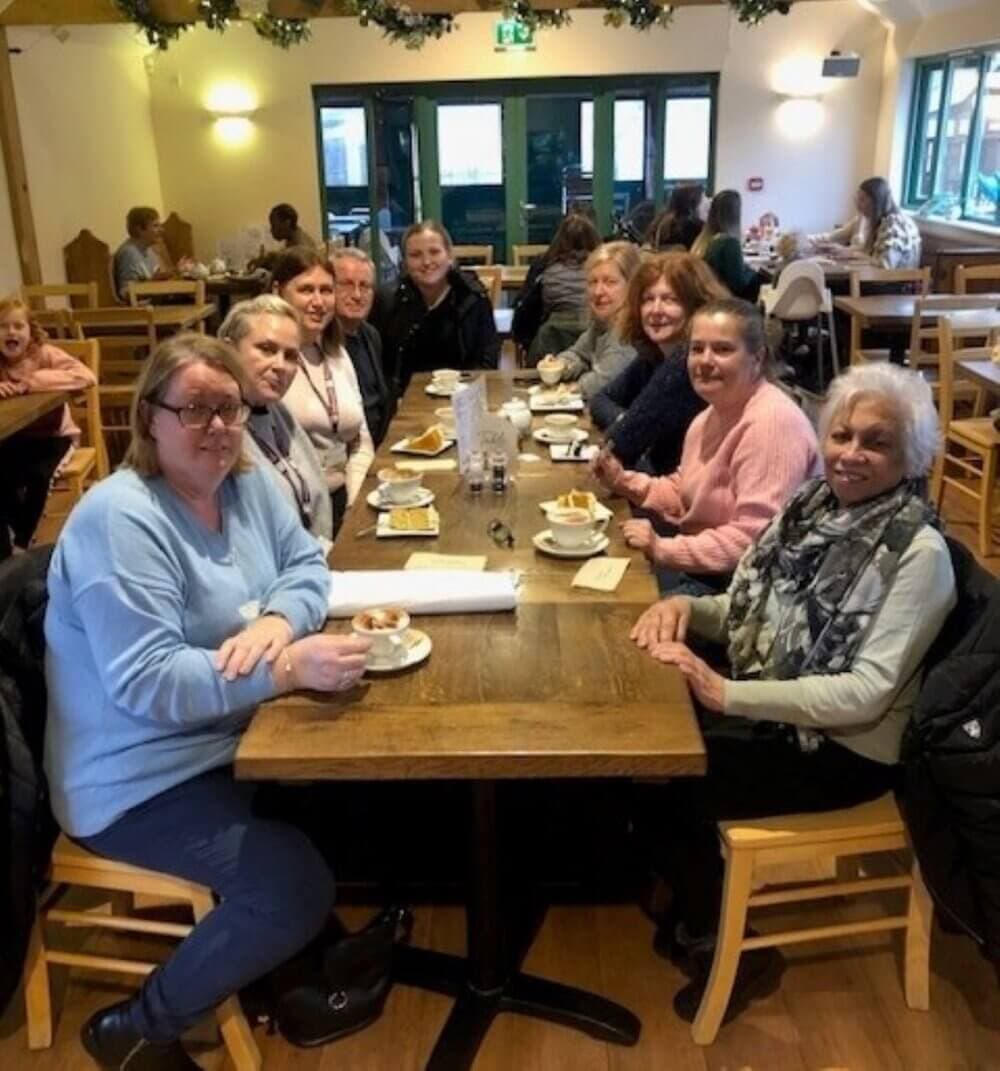 A group of women sitting around a long wooden table in a restaurant, smiling toward the camera. - Home Instead