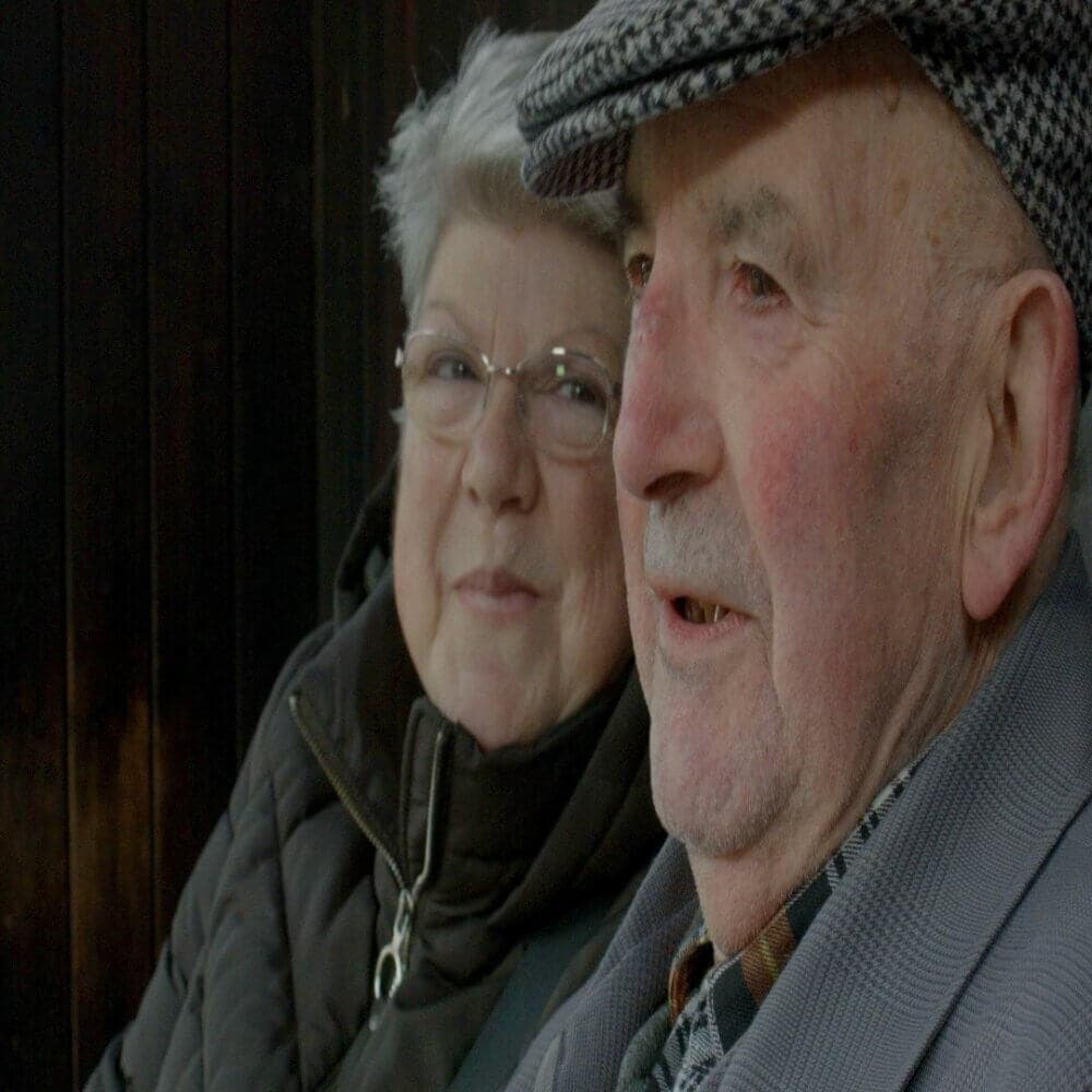 Elderly couple in coats and hats smiling warmly, with a dark background behind them. - Home Instead