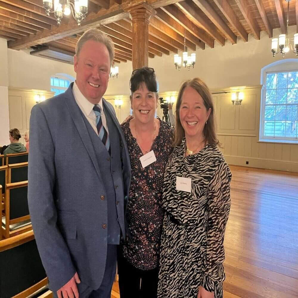 Three people standing and smiling in a well-lit room with wooden beams and windows in the background. - Home Instead