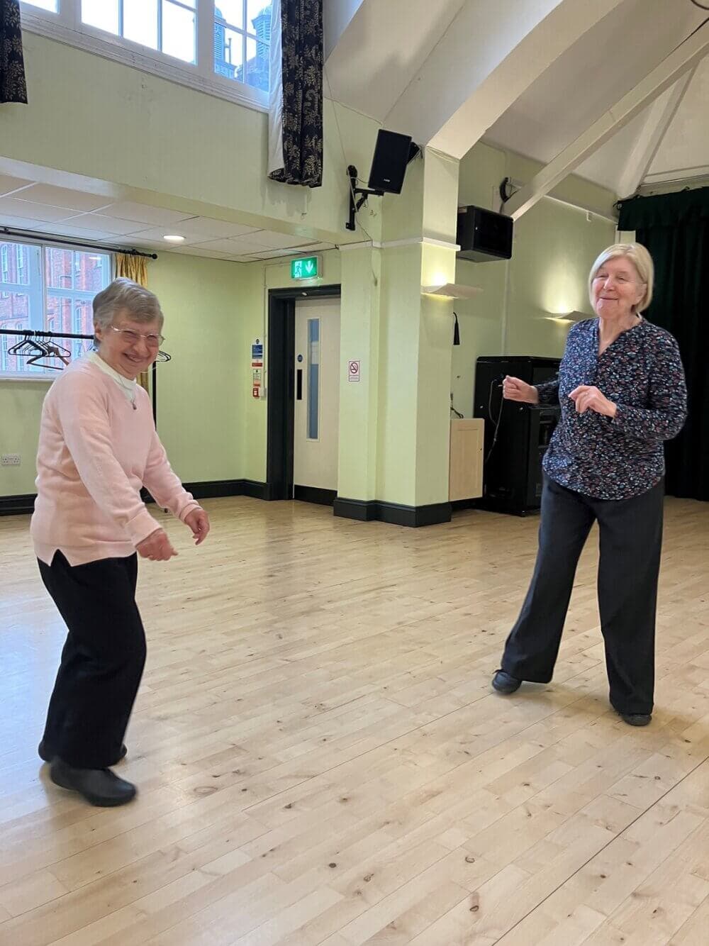 Two elderly women dancing in a brightly lit room with wooden floors, smiling and having fun. - Home Instead