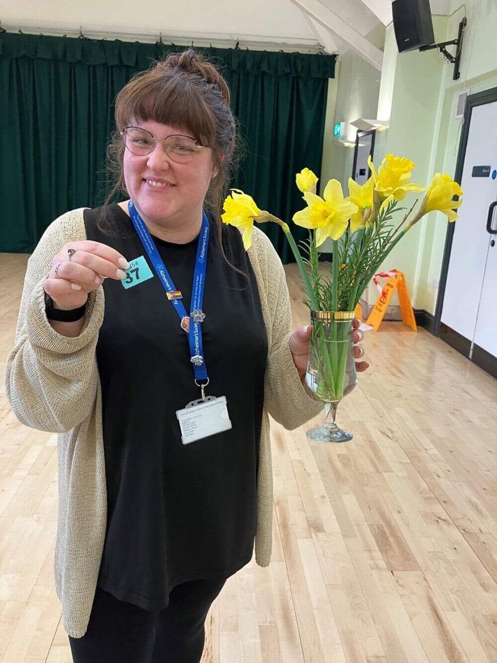 Smiling woman holding a number card and a vase of yellow daffodils in a large room with green curtains. - Home Instead