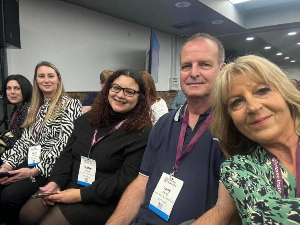 Group of people sitting together at an event, wearing name badges and smiling at the camera. - Home Instead
