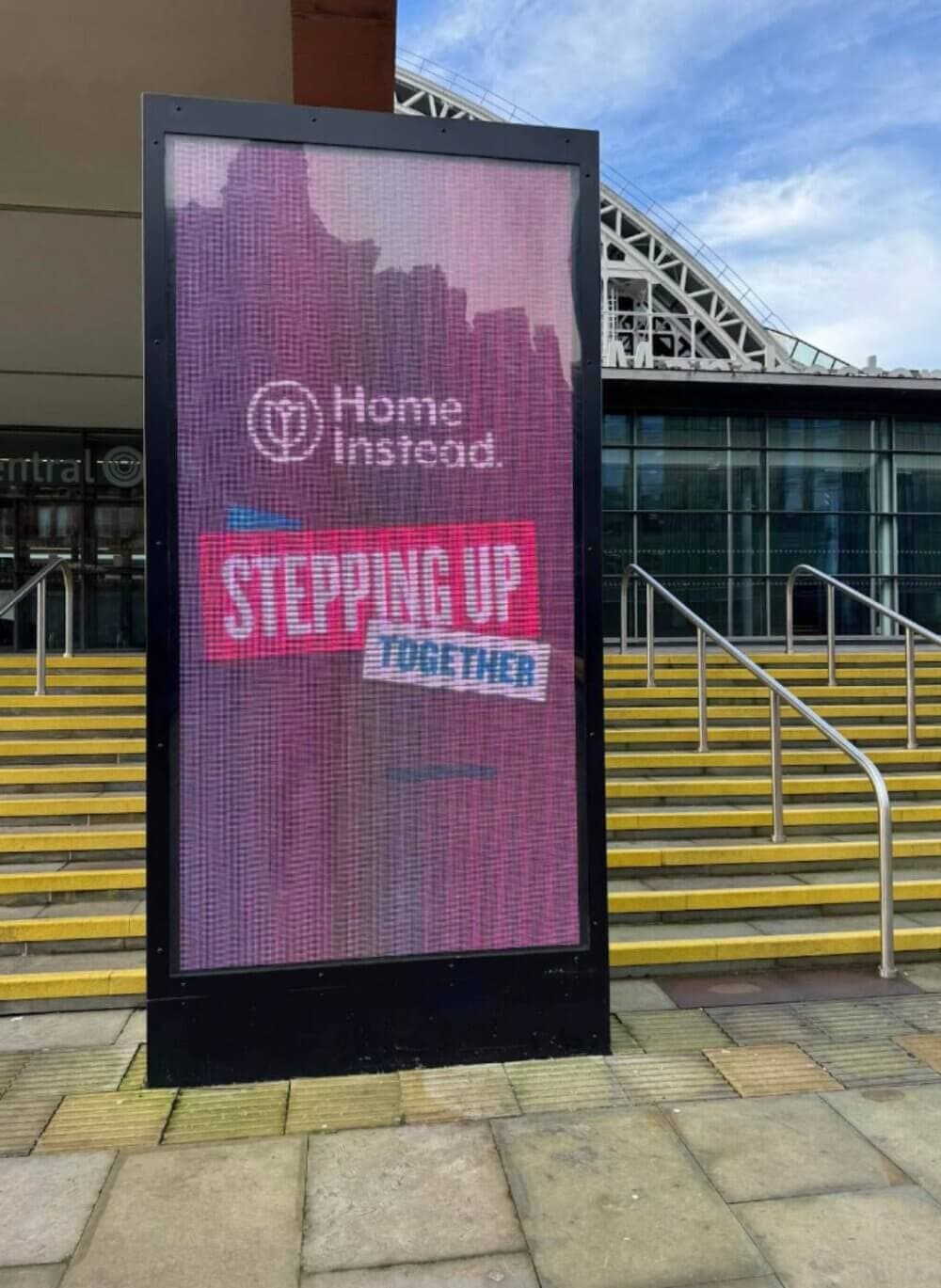 Outdoor electronic billboard reading "Home Instead STEPPING UP TOGETHER" in front of a building with stairs. - Home Instead