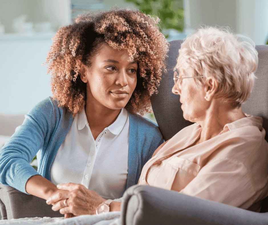 A young woman with curly hair sits and holds hands with an elderly woman in a chair, sharing a thoughtful moment. - Home Instead